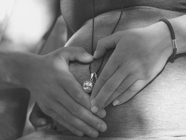 Gros plan sur le ventre d'une femme enceinte qui porte un bola. Elle et son compagnon posent leurs mains en forme de cœur autour du pendentif.