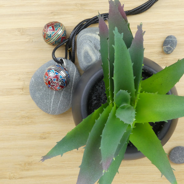 Bolas Acapulco dans leurs deux versions (argent et bronze) présentés dans un décor. Cailloux gris à rayures blanches et petite plante type cactus, le tout déposé sur un plateau en bois.