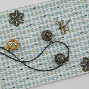 Bola Ecatepec dans ses 3 versions (bronze et noir, bronze et turquoise, doré) présentés dans un décor : déposés sur un tissu avec des motifs verts. Il y a aussi à proximité une libellule et deux fleurs couleur bronze.