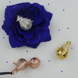 Les 3 versions du bola Goutte et cristaux présentés dans un décor. La version argent est dans une fleur en tissu bleue électrique. Le or rosé est ouvert de façon à voir les strass du dessus et ceux du fond et le or jaune fermé. Tous deux déposés à côté. Aucun ne contient de perle. Des petits strass bleus sont disséminés tout autour.