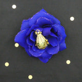 Version dorée (jaune) du bola Goutte et cristaux présentée dans un décor. Avec une perle verte clair insérée, il est dans une fleur en tissu bleue électrique.