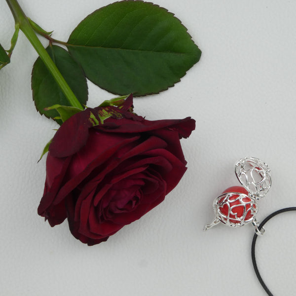 Bola Petits cœurs argent présenté sur un tissu blanc, ouvert et reposant sur une perle rouge. Une rose rouge est déposée à côté.