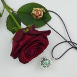 Bola Petits cœurs présenté dans ses deux versions sur un tissu blanc avec une rose rouge. Le bola doré avec perle rouge métallisé est déposé sur une feuille de la rose et le bola argent avec perle verte métallisée est déposé à côté de la rose.
