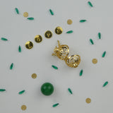 Bola de type cage doré présenté sous un autre angle. Il ne contient pas de perle et est posé sur un fond blanc. Des perles vertes et or sont disséminées tout autour en guise de décor. Une perle verte est également déposée à côte.