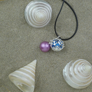 Bola arabesques couleur argent présenté sur du sable au milieu de coquillages nacrés. Une perle bleue y est insérée et une perle violette est posée à côté, seule