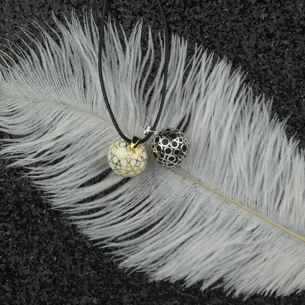 Les bola Bubbles dans leurs deux versions (dorée et argentée) présentés dans un décor : déposés sur une grande plume blanche elle même déposée sur un tissu pailleté noir.