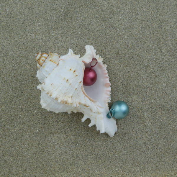 Bola Mini dans ses deux versions (bordeaux et bleu-vert) présenté dans un décor constitué de sable et où se trouve un gros coquillage: Le bola bordeaux est dans le coquillage et le bleu-vert à côté.